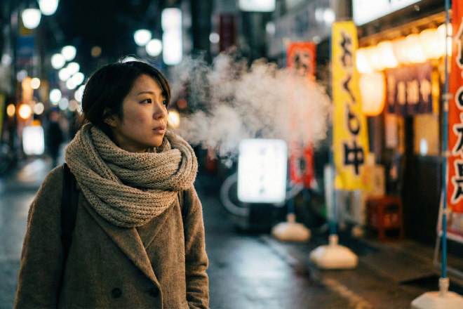 寒い冬の夜、マフラーをして歩く日本人女性。吐く息が白く、背景の店明かりに惹かれている。