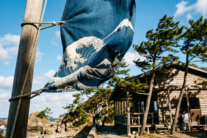 青空の下、強い風を受けてもしっかりと立っているのぼり旗。背景には日本の海岸沿いのカフェ。