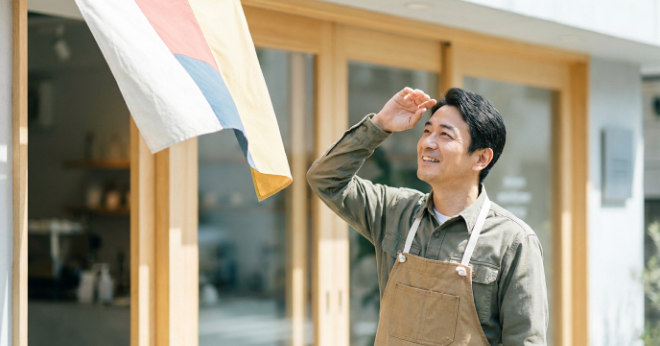 屋外で風にはためくのぼり旗を見上げ、生地の傷みがないか状態をチェックしている日本人の男性店長の写真